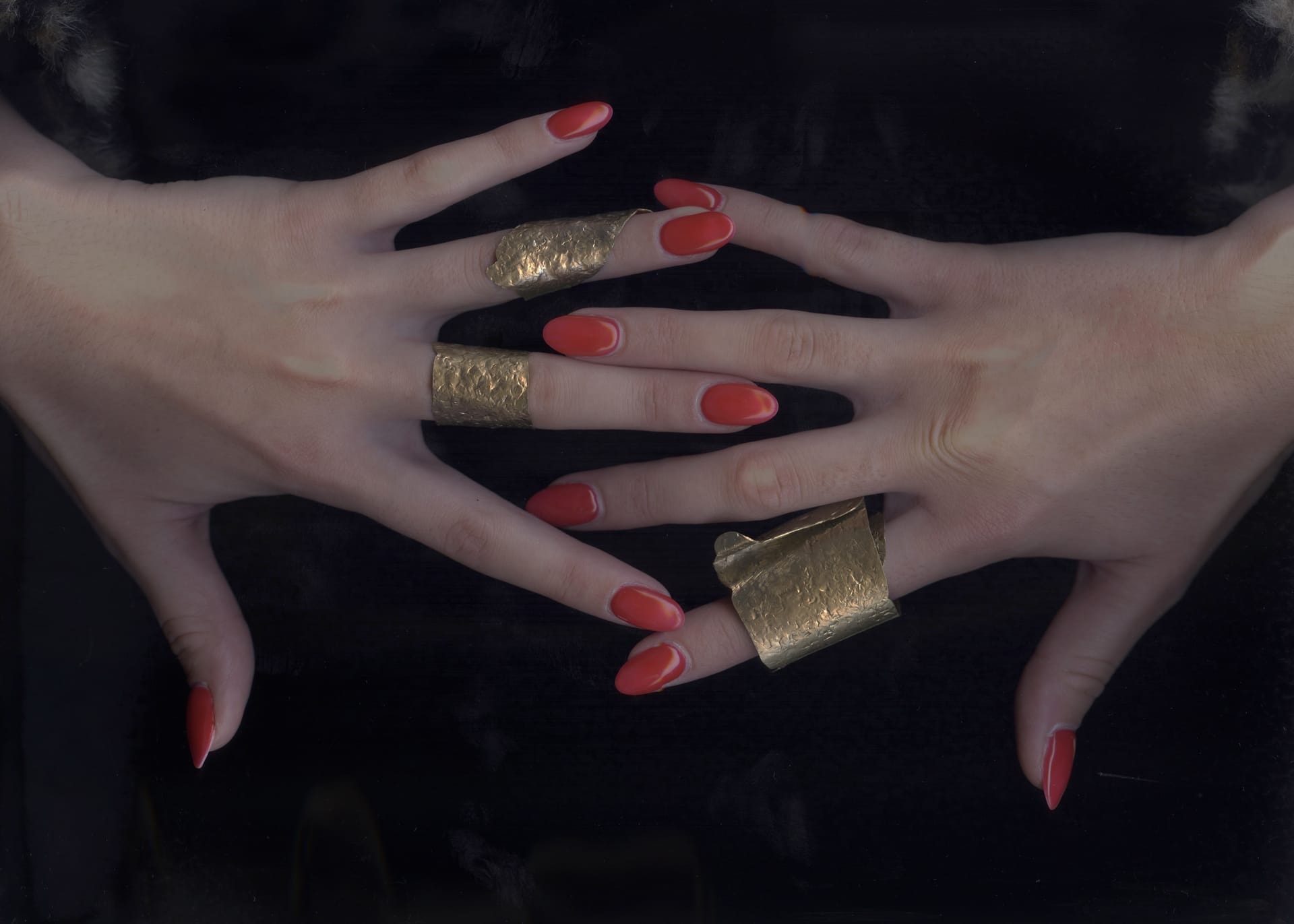 Hands wearing gold textured rings with red nails against a dark background