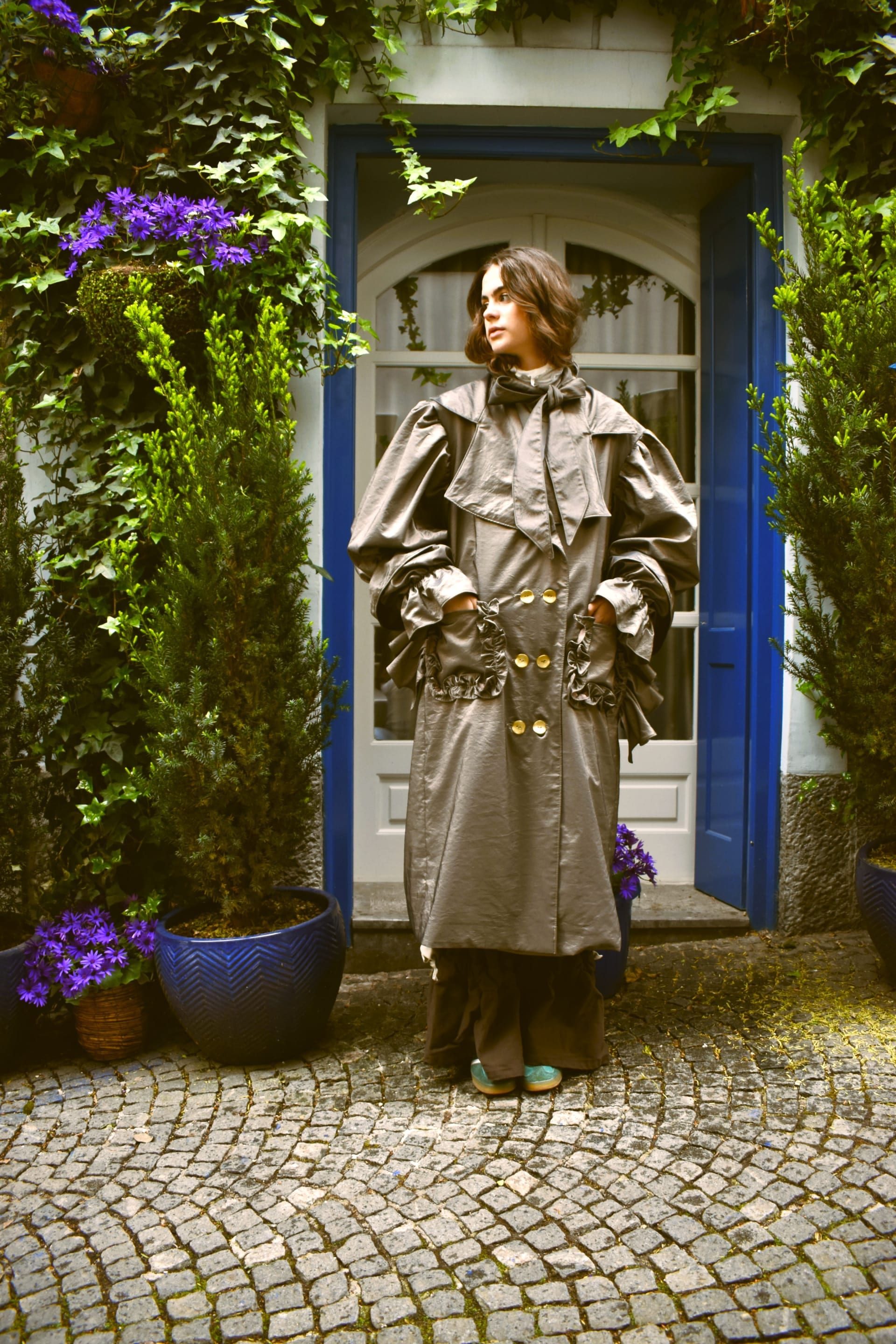 Model wearing a structured trench coat in front of a blue doorway with greenery